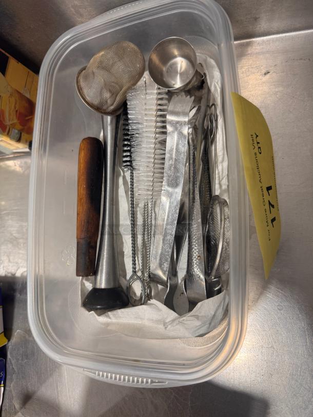 Assorted bar tools in plastic container, includes jigger, strainer, and muddler. Metal and wood materials, good condition.