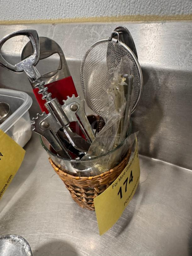 Lot of miscellaneous bar tools in a glass with a wicker holder, includes corkscrew and strainer, auction lot number 174.