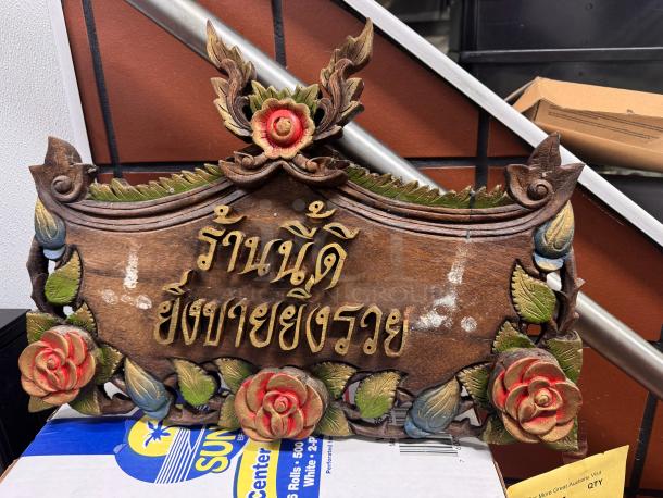Wooden Thai house sign with intricate carvings featuring floral motifs and gold lettering. Signs of wear add character.