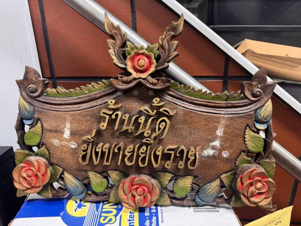 Hand-carved wooden Thai house sign with ornate floral design and Thai script, featuring red and green accents. Some wear visible.