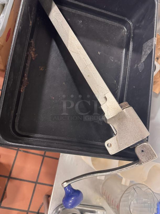 Heavy-duty industrial can opener with long metal handle, mounted on a black tray. Appears in used condition.