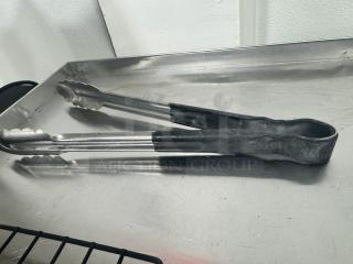 Pair of metal tongs with black grips on stainless steel surface. Used condition; suitable for kitchen or grilling use.