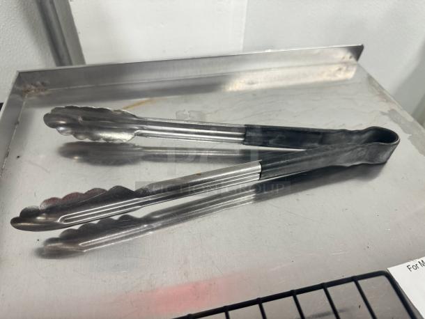 Lot of 2 metal tongs on a stainless steel surface, good condition. Handles feature a grip design for easy use.