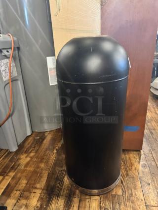 Black cylindrical trash can in used condition with a dome lid. Scuff marks visible on the surface.