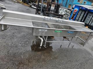 Three compartment stainless steel sink, 140", with dual drainboards, visible plumbing, used condition, commercial grade.