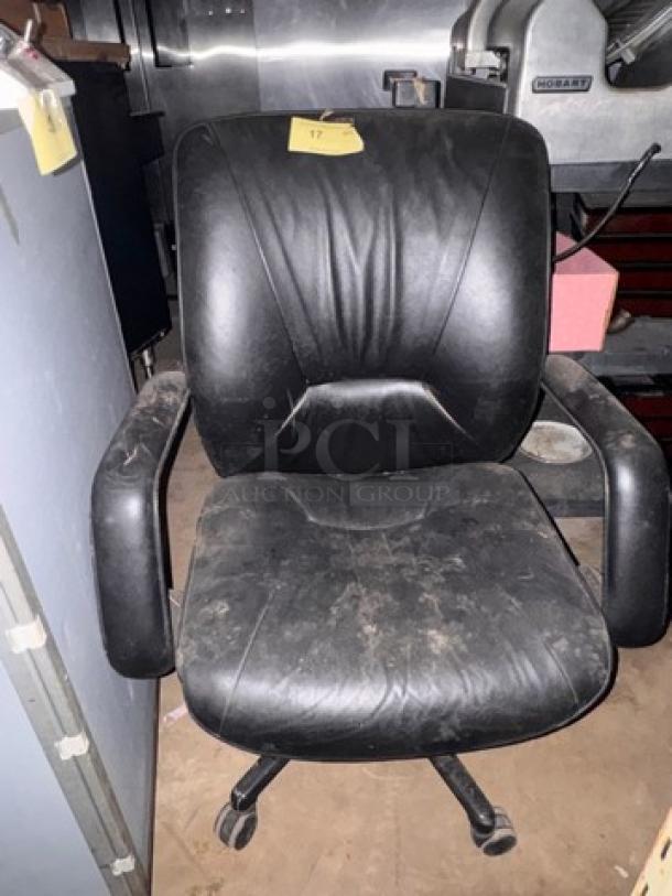 Black leather office chair with cushioned seat and backrest, armrests, on casters. Visible wear and dust on surface. Tag marked "17".