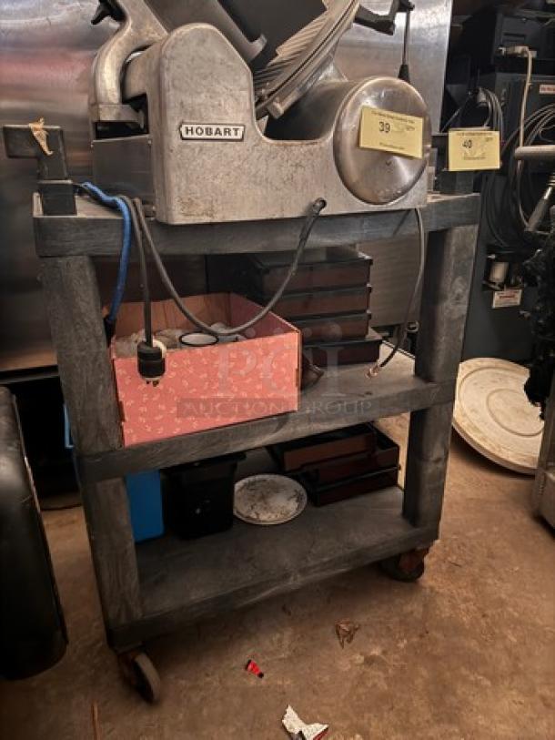 Gray utility cart with three shelves, caster wheels, and visible wear. Top shelf includes industrial slicer.