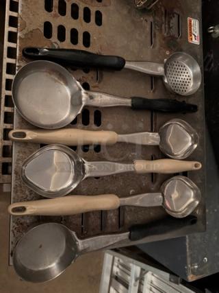 Set of food portion serving spoons on metal surface, various sizes, stainless steel with black and beige handles, used condition.