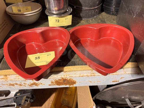 Red heart-shaped baking pans, set of two, on a shelf. Each pan includes a label with the auction site information and item number 55.