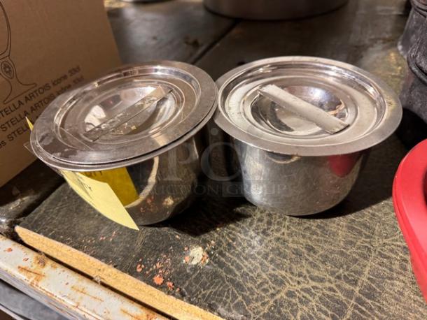 Two stainless steel containers with lids, labeled BIDX2, in used condition on a worn surface.
