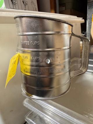 Metal flour/sugar sifter with volume markings, crank handle, and attached yellow tag. Good condition. Measures up to three cups.
