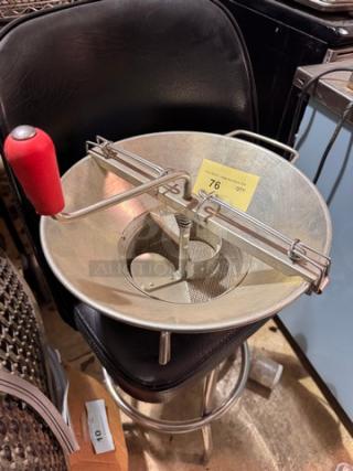 Stainless steel food mill with red handle, cranked mechanism. Visible tag indicating item number 76. In used condition.