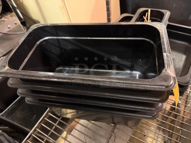 Stack of black plastic 1/3X6IN food pans, BIDX4 model, in good condition on a metal rack.