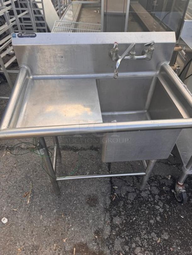 Stainless steel sink with side drainboard, wall-mounted faucet, in used condition. Ideal for kitchens or commercial use.