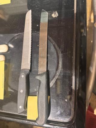 Lot of 2 knives with black handles on glass surface. One serrated blade, one straight. Visible engraved brand name on handle.