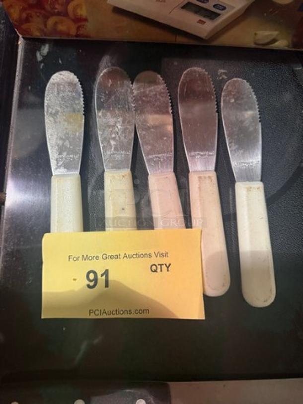 Lot of 5 spreader knives with white handles, serrated edges, used condition, auction tag number 91 visible.
