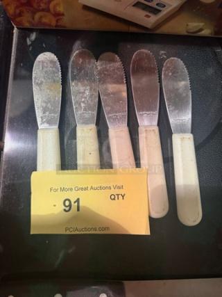 Lot of 5 spreader knives with white handles, serrated edges, used condition, auction tag number 91 visible.