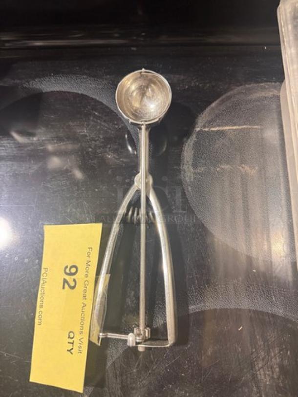 Stainless steel squeeze disher in new condition, featuring a round scoop and ergonomic handle. Auction tag reads "92".
