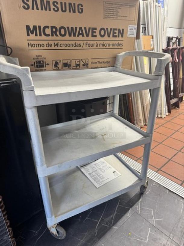 Gray utility cart with three shelves, plastic construction, some signs of wear, and four caster wheels for mobility.