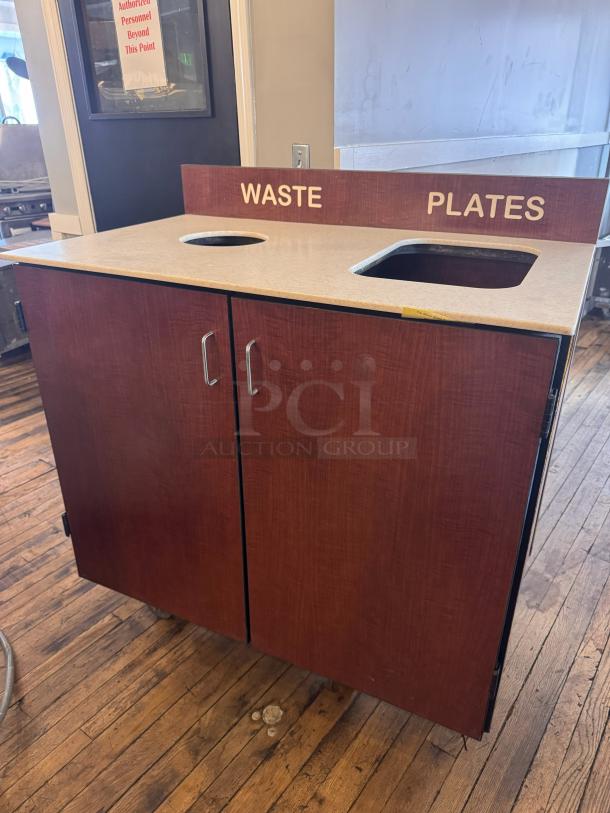 Wooden trash receptacle with labeled cutouts for waste and plates; dual doors below, smooth top surface, good condition.
