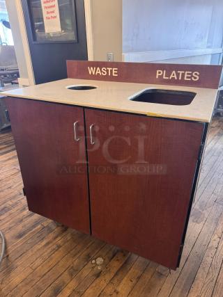 Wooden trash receptacle with labeled cutouts for waste and plates; dual doors below, smooth top surface, good condition.