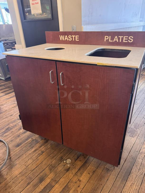 Wooden trash receptacle with labeled sections for waste and plates, two doors, and metal handles. Housing kitchen setting.