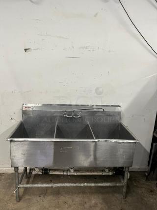 Stainless steel 3 compartment sink, used condition with visible wear. Includes faucet and drainage plumbing. Suitable for commercial use.