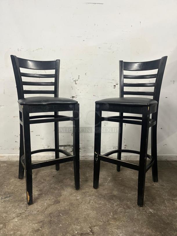 Two bar stools with black sandthru finish, featuring faux leather seats. Signs of wear on the legs.