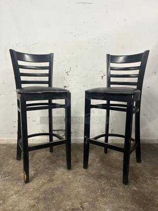 Two bar stools with black sandthru finish, featuring faux leather seats. Signs of wear on the legs.