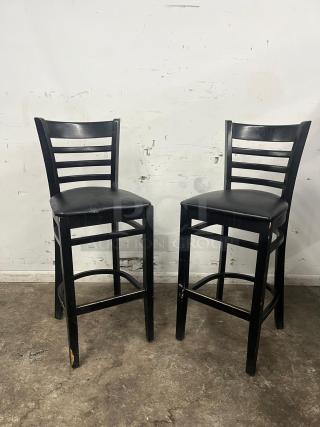 Black sandthru bar stools with faux leather seats. Features ladder-back design. Visible wear on legs and edges.