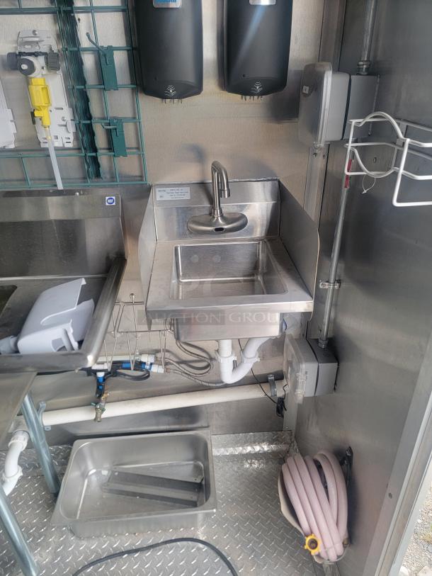Stainless steel sink area in a Rock Solid Cargo 24' mobile kitchen trailer, featuring plumbing and cleaning supplies.