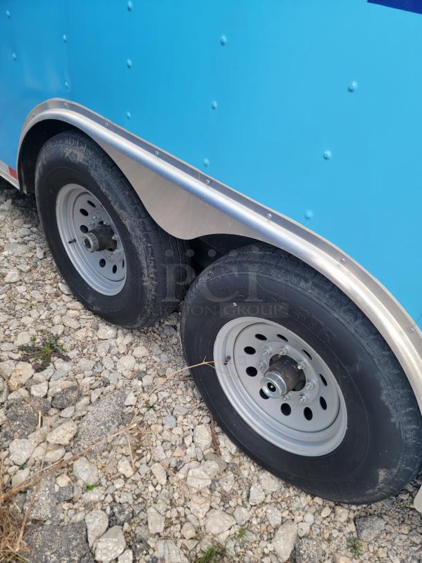 Dual-axle wheels of a 2020 24' Diamond Cargo mobile kitchen in blue, resting on gravel. Needs cleaning.