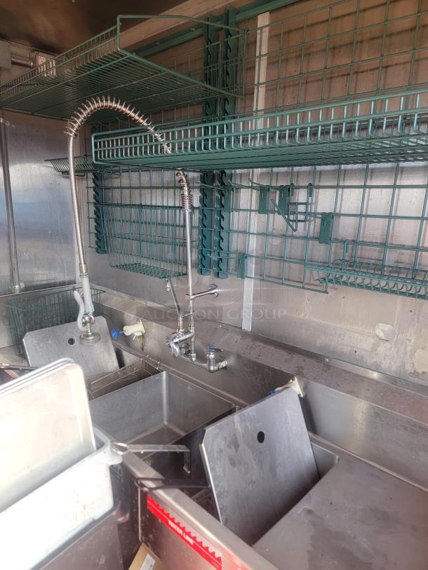 Stainless steel 3-compartment sink in a 2021 Titanium Cargo 28' Mobile Kitchen. Green wire shelving above; heavy-duty setup.