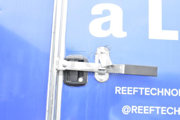 Blue 2021 Rock Solid Cargo trailer. Close-up of door latch. Markings read "REEF TECHNOLOGY @REEFTECHNOLOGY".