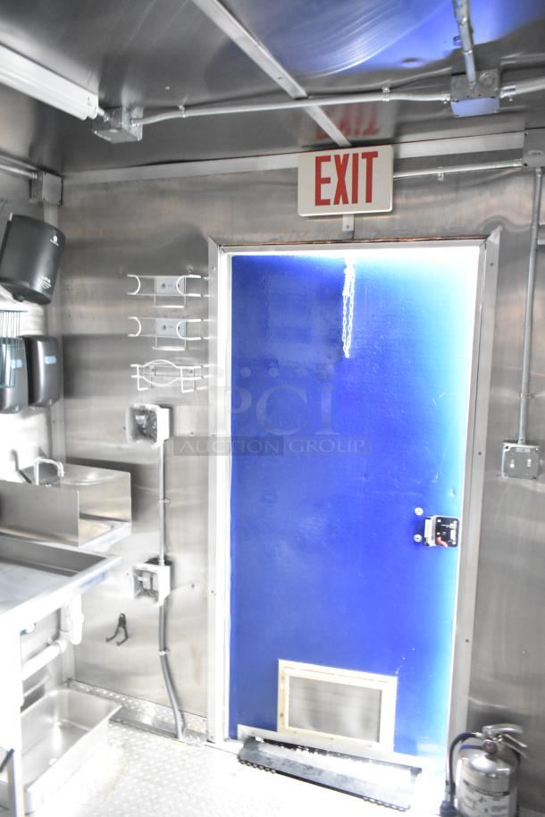 Interior of a brand new 2021 Rock Solid Cargo 24' mobile kitchen trailer. Features stainless steel surfaces, commercial sinks, blue exit door, and firewall with EXIT sign visible.