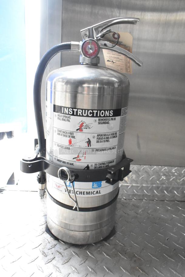 Fire extinguisher in mobile kitchen trailer, marked with safety instructions and mounted on a diamond plate floor.