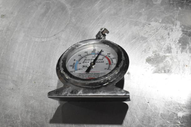 Thermometer gauge with metal casing on a stainless steel surface, displaying temperature range with "Refrigerator, Freezer, Dry Storage" labels.