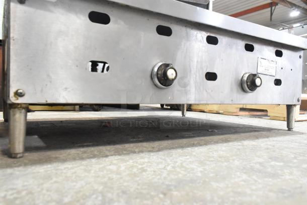 Commercial countertop gas-powered flat top griddle, stainless steel, dual control knobs, sturdy legs. Visible label indicates model details.