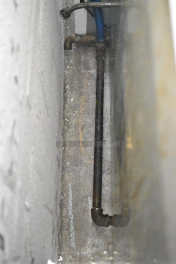 Image of a commercial floor-style pasta cooker, featuring metal piping and a blue hose, situated in an industrial setting.