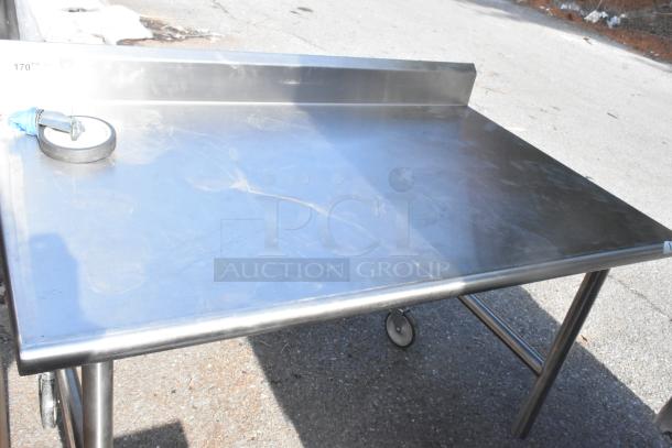 Stainless steel commercial table with wheels, used condition. Features a backsplash and sturdy legs; slight surface scratches.