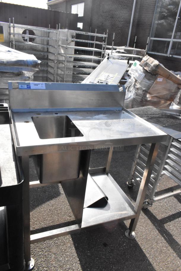 Stainless steel commercial single bay sink with integrated backsplash and undershelf. Slight wear visible. Auction lot tag on top.