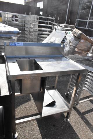 Stainless steel commercial single bay sink with integrated backsplash and undershelf. Slight wear visible. Auction lot tag on top.
