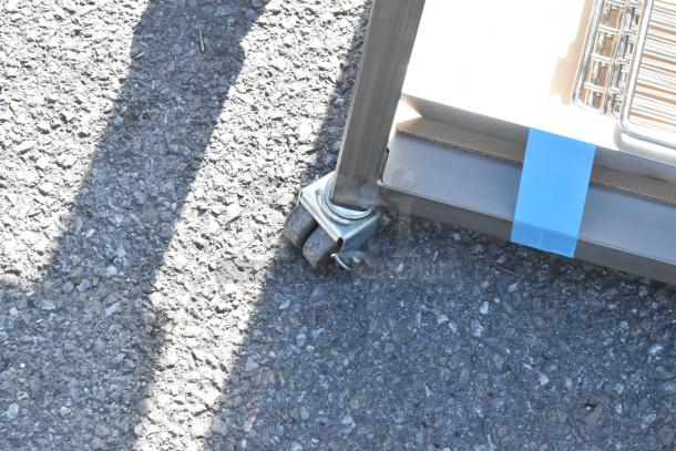 Commercial table with metal legs on sturdy casters, shown on asphalt. Features durable construction and mobility.