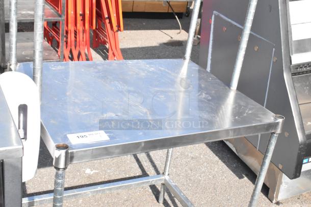 Stainless steel table with two-tier wire overshelf, slightly scuffed surface, and sturdy metal frame. Auction tag visible.