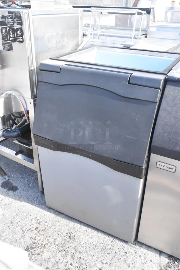 Commercial ice bin with black lid, stainless steel body, and "Ice-O-Matic" label. Appears in good condition.