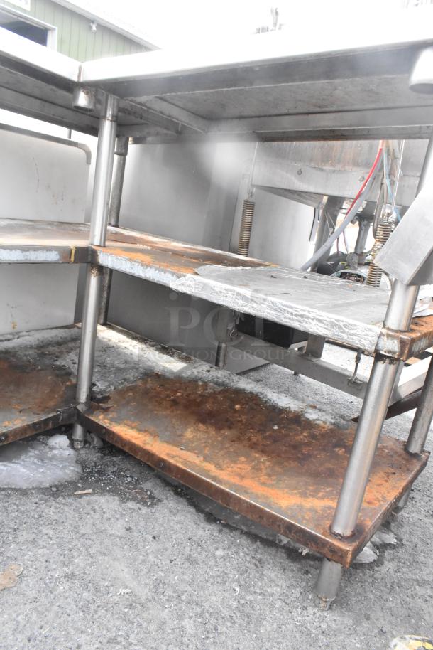 Commercial L-shaped table with two metal under shelves, showing signs of rust and wear. Ideal for industrial use.