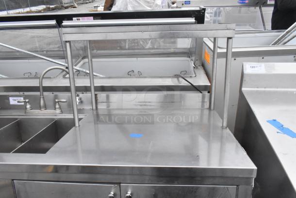 Commercial 3-bay stainless steel sink with over shelf and 3 doors, featuring dual faucets and storage space, in good condition.