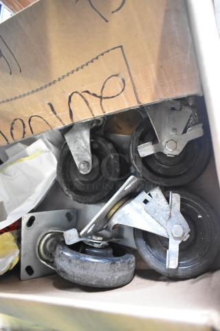 Lot of 4 commercial casters in a cardboard box, featuring metal frames and black rubber wheels, used condition.
