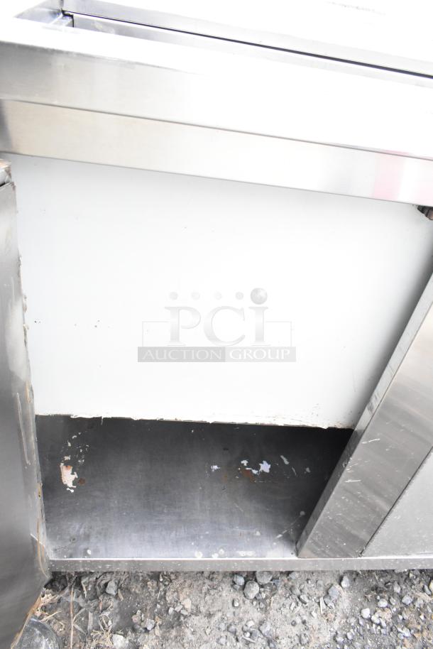 Stainless steel commercial counter with cut-out, over shelf, and three doors. Visible wear and minor rust spots on surface.