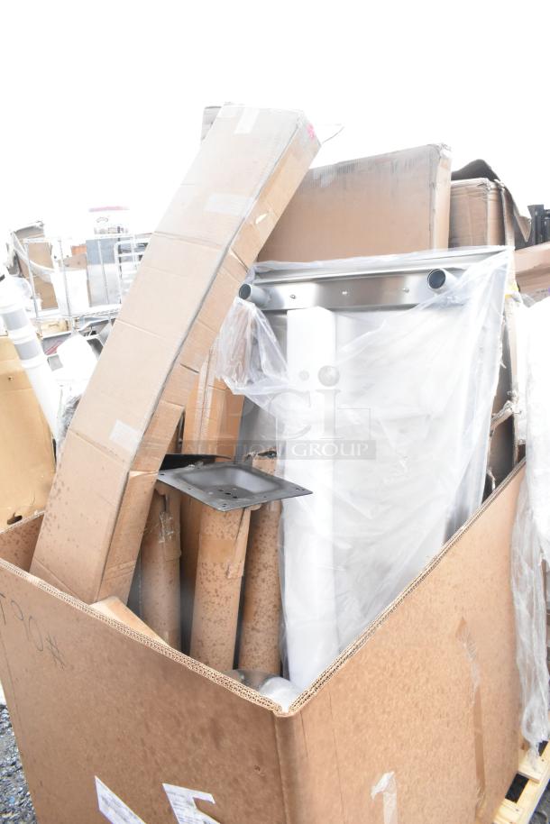 Cardboard box containing brand new scratch and dent items, including table and platform truck parts, some wrapped in plastic.